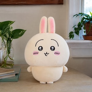 May include: A cream-colored plush rabbit toy with long ears featuring pink inner accents. The character has large black eyes, thin arched eyebrows, a small black mouth, and pink horizontal lines on its cheeks to represent a blushing expression.