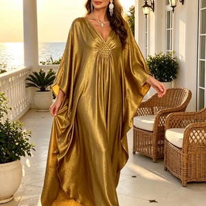 Op de afbeelding: Een vloerlange, metallic goudkleurige kaftanjurk met een diepe V-hals met een centraal geplooid detail, wijde, zwierige mouwen en een losjes gedrapeerd silhouet. De stof heeft een subtiele glinsterende textuur over het hele kledingstuk.