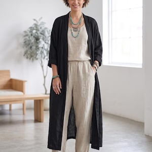 May include: A beige linen jumpsuit with a round neckline and elastic waistband paired with a long, open-front black duster coat. The outfit includes layered turquoise beaded necklaces, matching bracelets on both wrists, and simple white flat shoes.