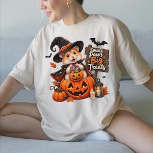 May include: A cream-colored t-shirt featuring a graphic of a hamster wearing a witch hat and cape, sitting inside a jack-o'-lantern while holding a candy bucket. The design includes a wooden sign that reads Small Paws Big Treats, bats, and a lantern.