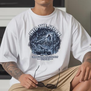 May include: A white t-shirt featuring a graphic print of a utility truck with a bucket lift and a worker repairing power lines during a snowstorm. The text reads INTO THE STORM, LINEMAN'S GRIND - NO WEATHER DELAY, and EST. [YEAR] in dark blue.