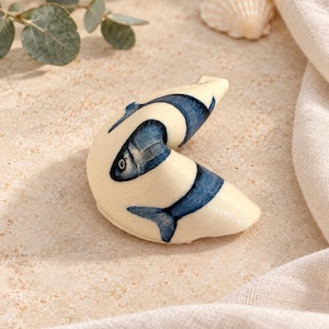 Può includere: Un oggetto a forma di biscotto della fortuna color crema con un'illustrazione a inchiostro blu di un pesce che si avvolge attorno alla sua superficie curva. Il disegno mostra la testa, il corpo e la coda del pesce in pigmento blu.