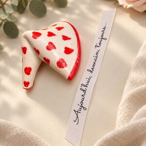 Puede incluir: Un adorno en forma de galleta de la fortuna de color crema decorado con pequeños corazones rojos y un borde rojo. Junto a él hay una tira de papel blanco con el texto cursivo negro "Aujourd'hui, demain, toujours..." impreso en el papel.