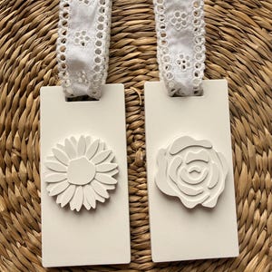 May include: Two rectangular white hanging tags with rounded corners. Each tag features a white floral relief emblem, one a daisy and one a rose. A white eyelet lace ribbon is looped through a slot at the top of each tag for hanging.