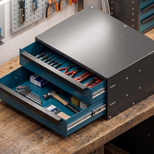 Può includere: Una cassetta degli attrezzi in metallo grigio scuro con due cassetti color ottanio aperti. Il cassetto superiore contiene varie pinze, mentre quello inferiore contiene un martello, un set di chiavi esagonali e delle piccole scatole.