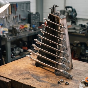 Op de afbeelding: Een metalen steeksleutelhouder met een driehoekig profiel, met een verticale stapel zilverkleurige steek-ringsleutels van verschillende groottes. De standaard heeft sleuven voor de sleutels en bevat kleine inbussleutels aan de bovenzijde.