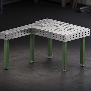 Pode incluir: Uma mesa de soldagem metálica em forma de L com um tampo prateado perfurado apresentando uma grade de orifícios circulares. A mesa é sustentada por cinco pernas verdes robustas com pés niveladores ajustáveis. Esquadros metálicos estão sobre o topo.