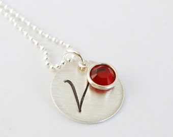 May include: A silver necklace with a round pendant featuring the letter "V" and a red gemstone.