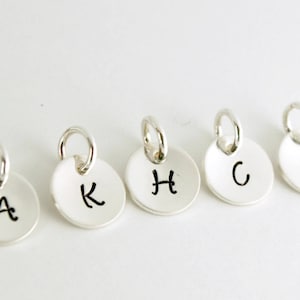 May include: Five silver charms with a round shape and a small loop at the top. Each charm has a different letter engraved on it: A, K, H, C, and B.