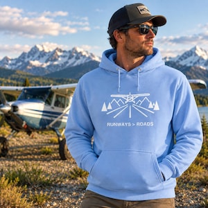 May include: A light blue hooded sweatshirt featuring a white graphic of an aeroplane landing on a runway with mountains and the text RUNWAYS > ROADS. The hoodie has a front kangaroo pocket, long sleeves, and matching drawstrings at the neckline.