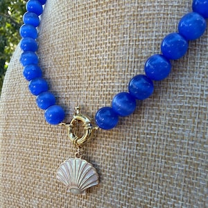 May include: A necklace featuring round, vibrant blue cat's eye beads strung together. The piece includes a gold-toned circular toggle clasp at the center, from which hangs a pale, iridescent seashell-shaped pendant with a smooth finish.
