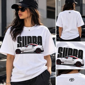 May include: A white short-sleeve t-shirt featuring a large black graphic of the word SUPRA above a white sports car with red accents. The back of the shirt displays a small black Toyota logo near the neckline, matching the logo on the cap.
