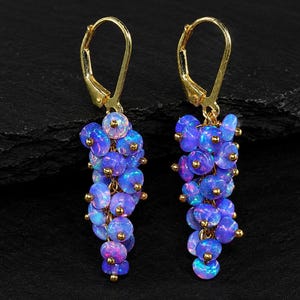 May include: A pair of gold leverback earrings featuring clusters of small, round, iridescent blue opal beads. Each earring is composed of multiple individual beads attached to a central gold chain, creating a dangling, grape-like shape.