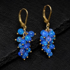 May include: A pair of gold-tone leverback earrings featuring clusters of small, round, iridescent blue opal-like beads. Each earring is composed of multiple spherical beads arranged in a grape-like bunch, suspended from a simple gold-coloured metal hook.