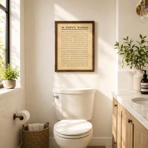 May include: A rectangular wall art print with a dark brown wooden frame featuring the title 50 POTTY WORDS in black text above a grid of letters on a beige background. The print is displayed on a white wall above a white porcelain toilet.