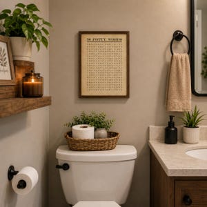 May include: A rectangular framed wall art piece with a dark wood frame featuring a word search puzzle titled 50 POTTY WORDS in black text on a beige background. The print is displayed on a neutral wall above a white toilet with a woven basket.