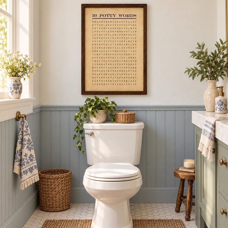 May include: A framed wall art print titled 50 POTTY WORDS featuring a word search puzzle in black text on a beige background. The print is displayed above a white toilet in a bathroom with blue wainscoting, a woven basket, and green plants.