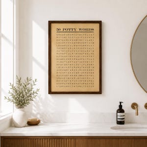 May include: A rectangular wall art print in a dark brown wooden frame featuring a word search puzzle titled 50 POTTY WORDS in black text on a tan background. The grid contains various letters arranged in rows and columns on a paper-like surface.