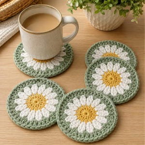 May include: A set of four round, crocheted coasters featuring a daisy pattern with a yellow centre, white petals, and a sage green border. One coaster supports a ceramic mug filled with a light brown beverage, while three others are arranged nearby.