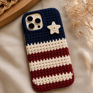 May include: A rectangular smartphone case featuring a crocheted cover with a patriotic design. The top section is dark blue with a single cream-colored crocheted star, while the lower portion consists of alternating horizontal stripes of cream and red.