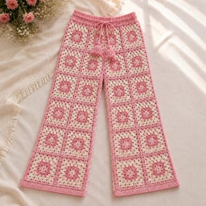 Könnte beinhalten: Häkelhose mit einem sich wiederholenden Granny-Square-Muster in abwechselnden Farbtönen von Hellrosa und Creme. Die Hose hat einen weiten Beinschnitt, einen hoch sitzenden elastischen Bund und einen passenden rosa Kordelzug mit zwei Quasten.