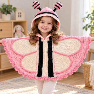 May include: A crochet hooded poncho featuring a pink and cream butterfly wing design with a black central stripe and two antennae on the hood. The garment has a scalloped pink border and is constructed with a textured yarn stitch pattern throughout.