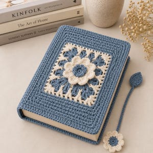 May include: A rectangular book cover made of blue and cream-colored yarn, featuring a central crocheted flower motif surrounded by a square pattern. A matching blue crocheted bookmark with a leaf shape and a small flower at the end is attached.