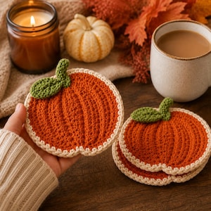 May include: A set of round, orange crochet coasters shaped like pumpkins, each featuring a textured ribbed design, a small green leaf accent at the top, and a cream-coloured border. The coasters are crafted with yarn and have a flat, circular form.