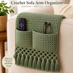 Könnte beinhalten: Ein häkeln Sofa-Armlehnen-Organizer in Salbeigrün mit zwei Taschen, einer dekorativen Gitterfranse mit Quasten sowie dem Text "Crochet Sofa Arm Organizer" und "KEEP YOUR ESSENTIALS CLOSE & ORGANIZED". Eine Fernbedienung und eine Brille stecken darin.