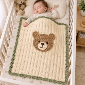 Può includere: Una coperta per neonati lavorata all'uncinetto color crema con un'applicazione centrale a forma di muso d'orso marrone e un bordo smerlato verde salvia. La coperta ha una consistenza a coste ed è rettangolare, con cuciture dettagliate sul muso.