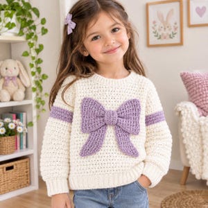 May include: A long-sleeved cream-colored crochet sweater featuring a large, textured lavender bow centered on the front and a matching lavender horizontal stripe across the chest and sleeves. The sweater has a ribbed crew neckline and cuffs.
