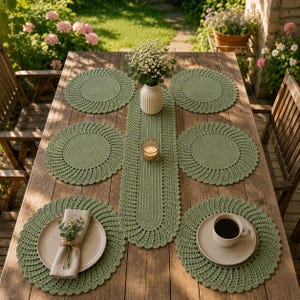 May include: A sage green crochet table setting set including a long runner and six round placemats. The runner features a central candle in a glass holder and a white flower vase. One placemat holds a beige napkin, another a ceramic coffee mug.