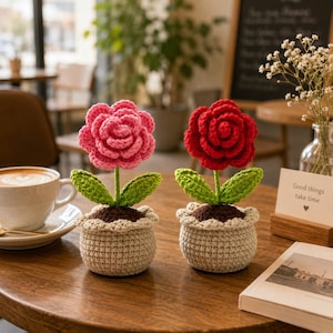 Op de afbeelding: Twee gehaakte bloemen in pot, een met een roze roos en een met een rode roos, elk met groene bladeren en donkerbruine aarde, in beige gestructureerde potten met een geschulpte bovenrand, gemaakt van garen met zichtbare stekenpatronen.