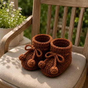 May include: A pair of dark brown crocheted booties featuring a high-top ankle design, a thick textured knit pattern, and decorative front laces that end in small, round pom-poms. The booties are constructed with a sturdy base and a ribbed cuff.