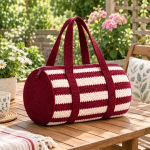 Peut inclure: Un sac cylindrique en crochet avec des rayures horizontales bordeaux foncé et blanc cassé. Le sac possède deux anses en crochet bordeaux assorties et une fermeture éclair sur le dessus. Le panneau latéral circulaire présente un motif en spirale.