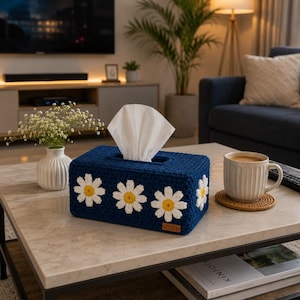 May include: A rectangular tissue box cover made of dark blue crocheted yarn featuring white daisy motifs with yellow centers. The cover has a rectangular opening at the top for dispensing white tissues and a small brown rectangular label on the side.
