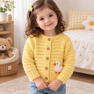 May include: A bright yellow long-sleeved crochet cardigan featuring a round neckline, five brown wooden buttons down the front, and a small white duck appliqué with an orange beak and feet stitched onto the lower left side of the garment.