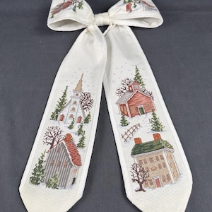 May include: A cream-colored fabric bow featuring cross-stitched winter scenes. The two long tails display a white church, a grey house, a red schoolhouse, and a tan house, all surrounded by green pine trees, bare brown trees, and white snow.