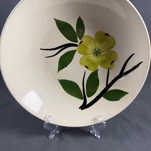 May include: A round, cream-colored ceramic bowl featuring a hand-painted design of a single yellow dogwood flower with green leaves on a black branch. The interior surface of the bowl is smooth and glossy, showcasing the floral motif centered.