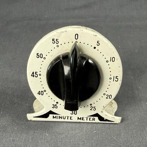 May include: A vintage Lux Minute Meter kitchen timer with a cream-colored metal body and a large black central control knob. The circular face features black numerical markings from 0 to 55, with the text "MINUTE METER" printed in black at the base.