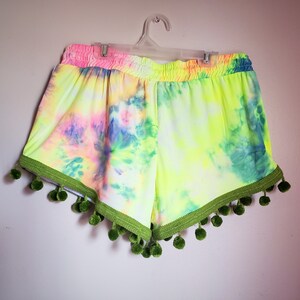 K&ouml;nnte beinhalten: Batik-Shorts in Neongelb, Rosa und Blau, mit gr&uuml;nen Pompons verziert. Der elastische Bund ist rosa. Diese Shorts h&auml;ngen an einem wei&szlig;en Kleiderb&uuml;gel vor einer wei&szlig;en Wand. Die Shorts sind ein lebendiges und farbenfrohes Kleidungsst&uuml;ck.