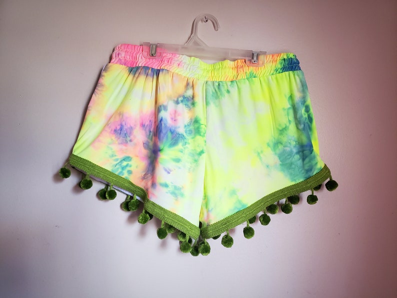 K&ouml;nnte beinhalten: Batik-Shorts in Neongelb, Rosa und Blau, mit gr&uuml;nen Pompons verziert. Der elastische Bund ist rosa. Diese Shorts h&auml;ngen an einem wei&szlig;en Kleiderb&uuml;gel vor einer wei&szlig;en Wand. Die Shorts sind ein lebendiges und farbenfrohes Kleidungsst&uuml;ck.