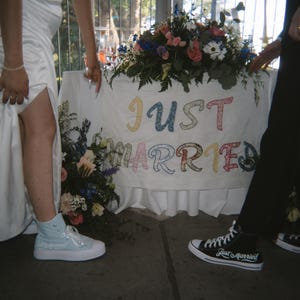 May include: A white fabric banner with the text JUST MARRIED in colorful floral-patterned letters, displayed on a table with floral arrangements. One light blue high-top sneaker and one black high-top sneaker with white text are visible.