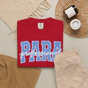 May include: A red short-sleeved t-shirt featuring the word PARA in large blue block letters with white polka dots, and the word professional in white cursive script layered over it. The shirt has a Comfort Colors brand label at the inner neckline.
