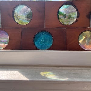 May include: Five rectangular wooden blocks, each featuring a circular inset window. These windows display miniature painted scenes, including landscapes with trees, a body of water, a starry night sky, and a coastal view with a lighthouse.