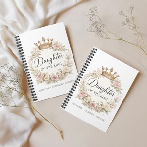 May include: Two spiral-bound notebooks with white covers featuring a gold crown illustration atop a floral wreath of pink and cream roses. The text reads Daughter OF THE KING with the words BELOVED ADORNED CHOSEN printed below the floral design.