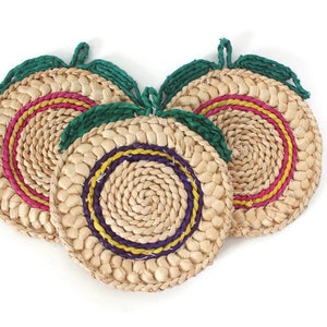 May include: Three woven straw trivets in the shape of a peach with green leaves. The trivets have a pink, yellow, and purple trim.