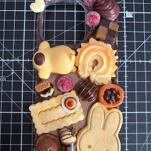Puede incluir: Una funda para teléfono decorada con dijes en miniatura con temática de comida, que incluyen un personaje amarillo, una galleta con forma de conejo, una galleta Lotus, trufas de chocolate, bayas rojas y un pequeño vaso de bebida.