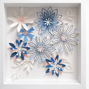 Peut inclure: Un cadre carré blanc de type boîte contenant des fleurs en papier superposées dans des tons de bleu, blanc et rose pâle. La composition présente plusieurs fleurs à pétales multiples et des feuilles en papier délicates sur fond blanc.