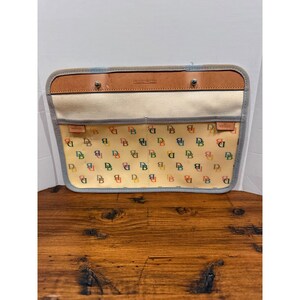 May include: A rectangular Dooney & Bourke pouch featuring a cream-colored canvas body with a repeating multicolored DB monogram print. The pouch includes a tan leather top trim with two silver-tone metal studs and a light gray fabric border.